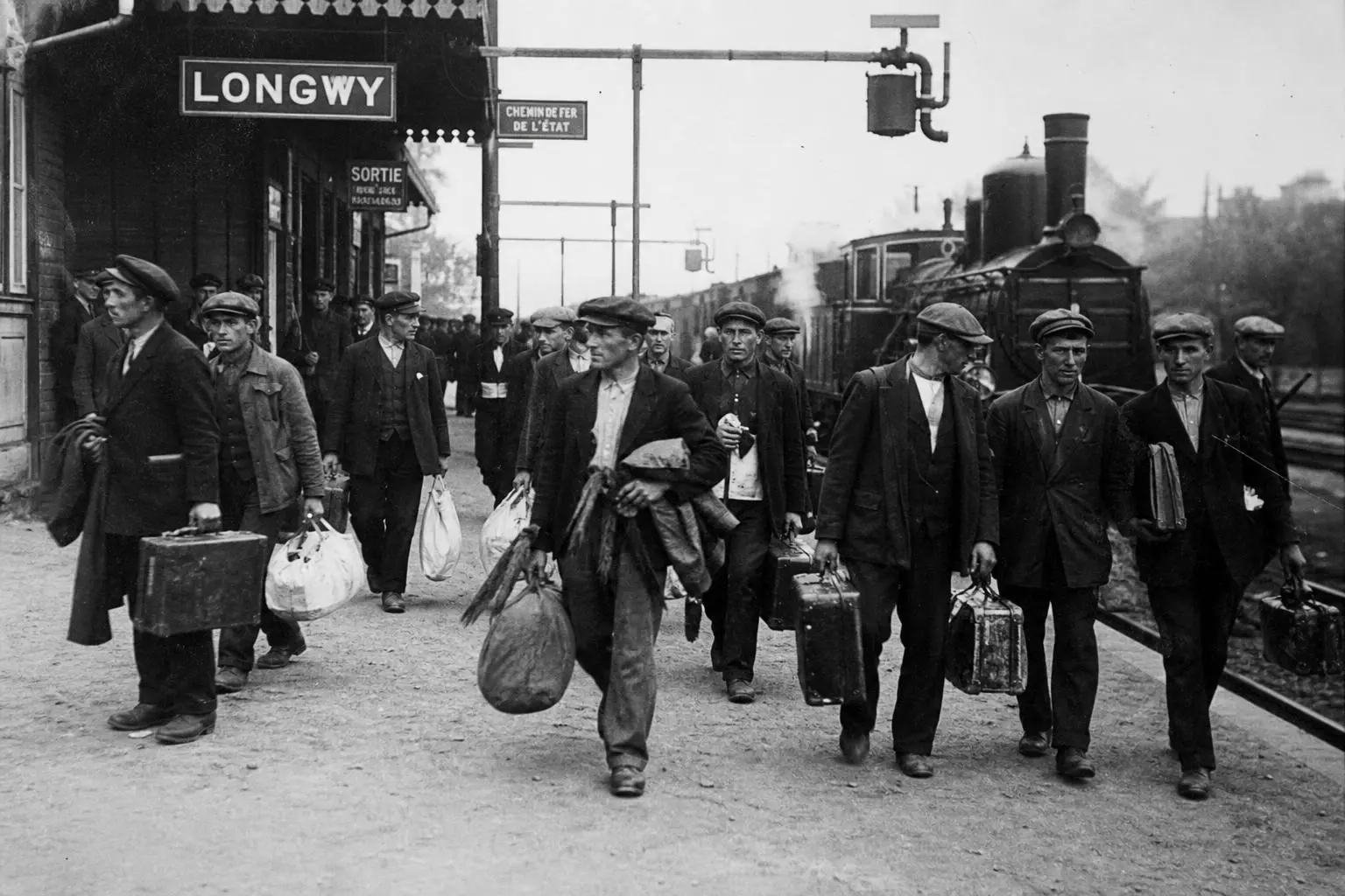 Travailleurs migrants sortant d’une gare, chargés de valises et de sacs, se dirigeant vers le quai, en noir et blanc.